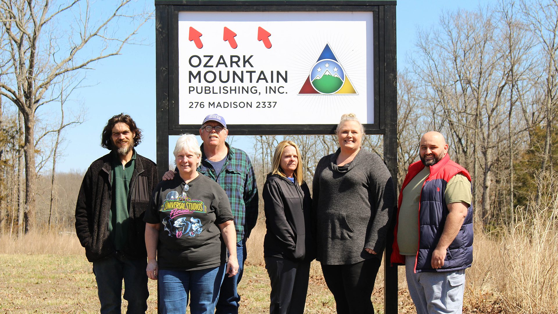 Group photo of three men and three women in front of an Ozark Mountain Publishing, Inc. signage