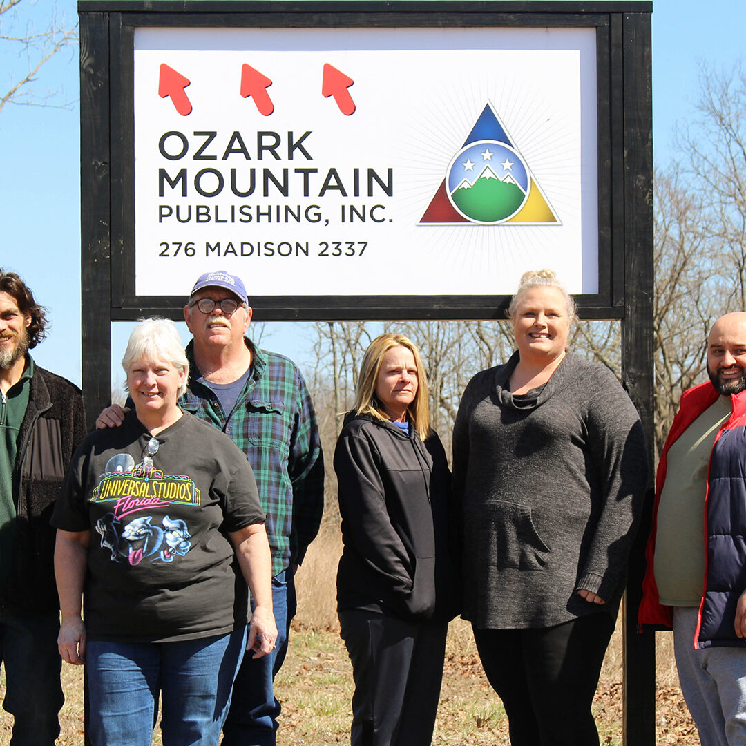 Group photo of three men and three women in front of an Ozark Mountain Publishing, Inc. signage